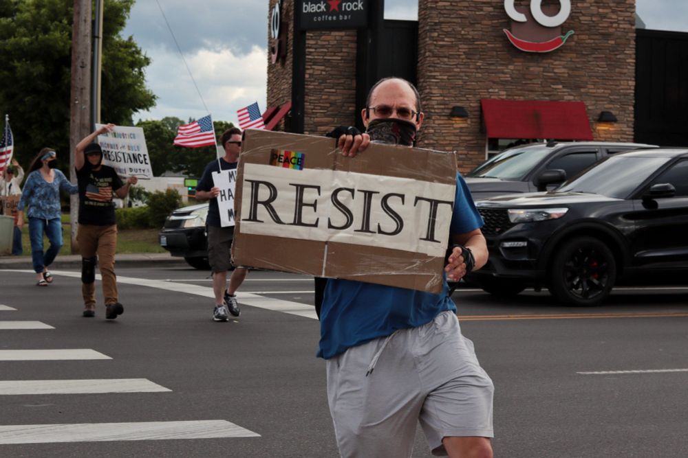 A protestor jogs across the street towards the viewer with a picket sign. The sign reads: “RESIST”. A rainbow-patterned sticker adhered to the left side of the sign reads: “PEACE”. Other protestors walk across the street in the background. 

At 5:00pm on May 7th, 2025, protestors gathered at the intersection of NE Fourth Plain Blvd and NE Andresen Rd outside the Vancouver, WA, Tesla dealership to protest the fascist regime of President Donald Trump and unelected billionaire Elon Musk.
