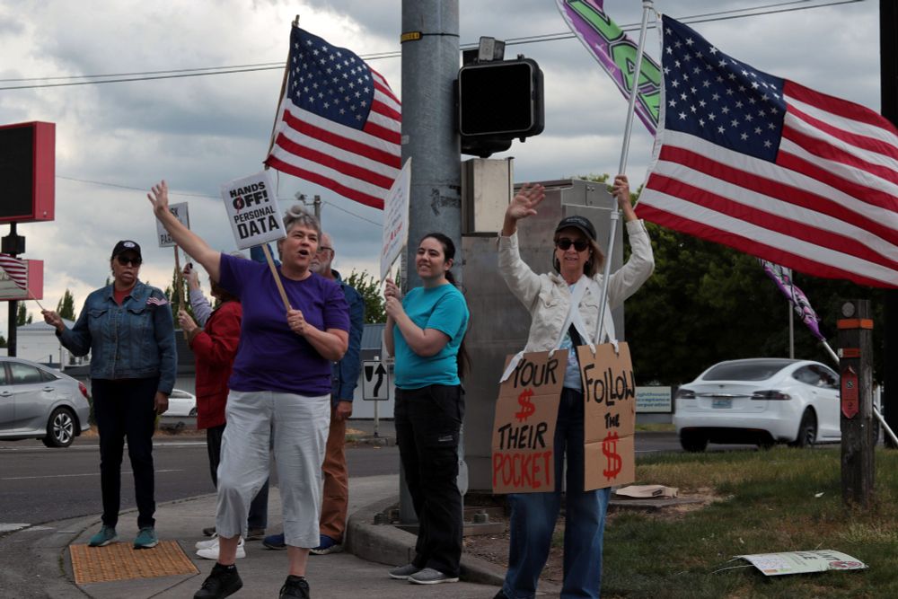 A group of protestors stand at the corner of the sidewalk. The protestors wave at passing cars, some holding full-sized US flags and others grasping picket signs. From left to right, the picket signs read: “HANDS OFF! PERSONAL DATA” and “YOUR $ THEIR POCKET | FOLLOW THE $”. 

At 5:00pm on May 7th, 2025, protestors gathered at the intersection of NE Fourth Plain Blvd and NE Andresen Rd outside the Vancouver, WA, Tesla dealership to protest the fascist regime of President Donald Trump and unelected billionaire Elon Musk.
