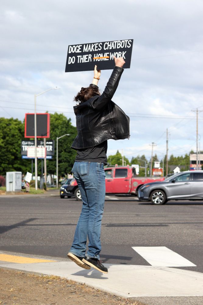 A protestor at the edge of the sidewalk with their back to the viewer holds a sign above their head. The visible side of the sign reads: “DOGE MAKES CHATBOTS DO THEIR HOMEWORK”. A red line is striked through the word, “HOME”. 

At 5:00pm on May 14th, 2025, protestors gathered outside the Vancouver, WA Tesla dealership to protest the fascist regime of US President Donald Trump and unelected billionaire oligarch Elon Musk. 
