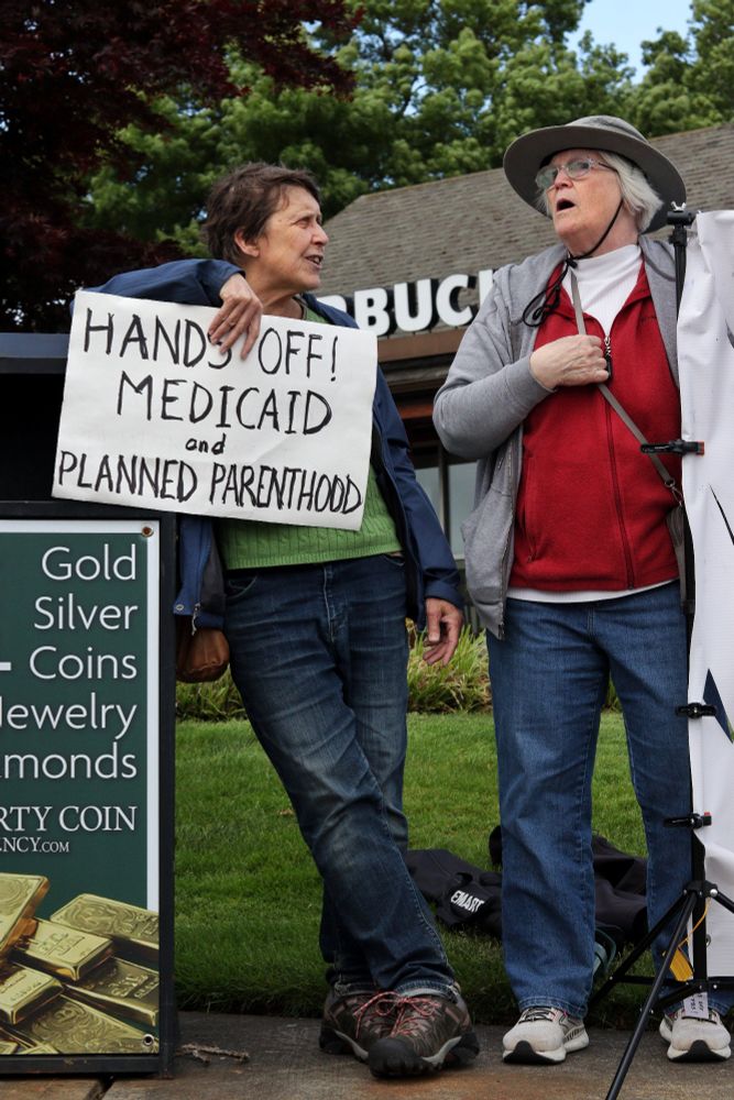 A protestor leans against a trash can while holding up a sign which reads, "HANDS OFF! MEDICAID and PLANNED PARENTHOOD." The first protestor speaks to another protestor who holds up one end of a banner.

At 4pm on May 19th, 2025, demonstrators in Vancouver, WA gathered at the intersection of Mill Plain and Chaklov to protest the destruction of essential social services by US President Donald Trump's fascist regime.