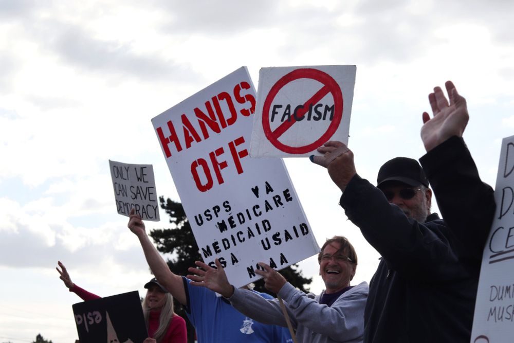 Several protestors hold picket signs above their heads for passing cars to read. The signs on the left side of the image read: “ONLY WE CAN SAVE DEMOCRACY” and “HANDS OFF | USPS | VA | MEDICARE | SSA | USAID | NOAA”. The sign on the right side of the image depicts the word, “FACISM”, crossed out in a red circle.

At 5:00pm on May 14th, 2025, protestors gathered outside the Vancouver, WA Tesla dealership to protest the fascist regime of US President Donald Trump and unelected billionaire oligarch Elon Musk. 
