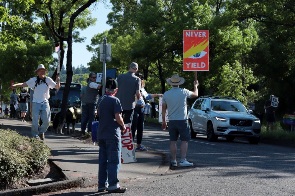 On the left side of the image, a group of protestors stand in a line along the sidewalk. The protestors face away from the viewer, looking into oncoming traffic. On the right side of the image, a car drives toward the viewer. The protestos hold picket signs. The sign in the center of the image depicts a close-up illustration of President Donald Trump's eye and eyebrow. The sign reads: “NEVER YIELD”. 

At 3:45pm on May 5th, 2025, protestors gathered outside the Portland Tesla dealership on Macadam Ave to protest the fascist regime of Donald Trump and Elon Musk
