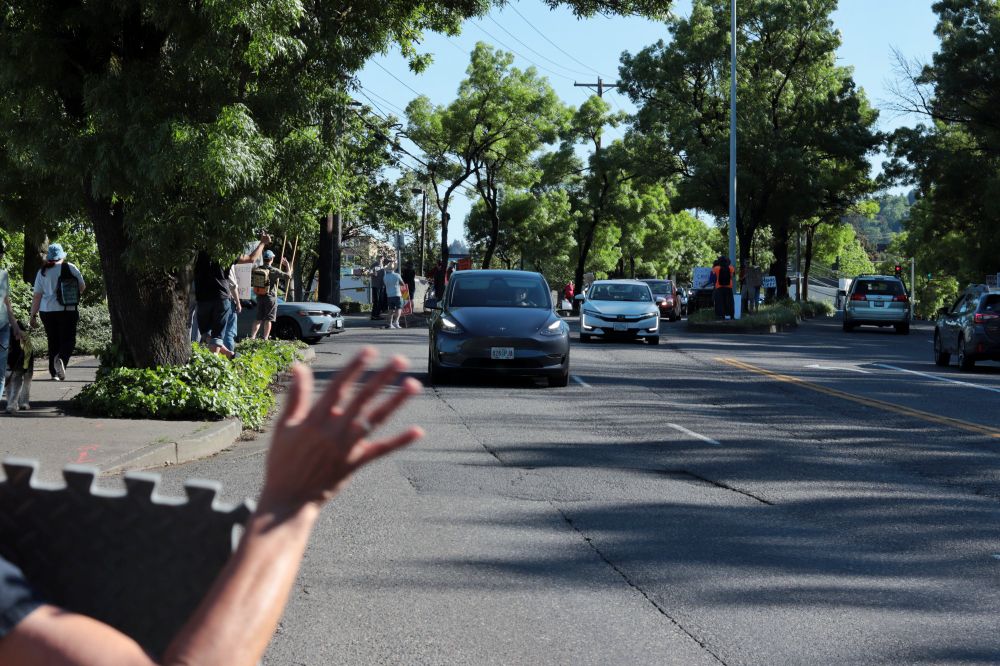 A grey Tesla vehicle drives towards the viewer. In the foreground, a protestor is waving towards traffic. I. The background, many more protestors line the road.

At 3:45pm on May 5th, 2025, protestors gathered outside the Portland Tesla dealership on Macadam Ave to protest the fascist regime of Donald Trump and Elon Musk.
