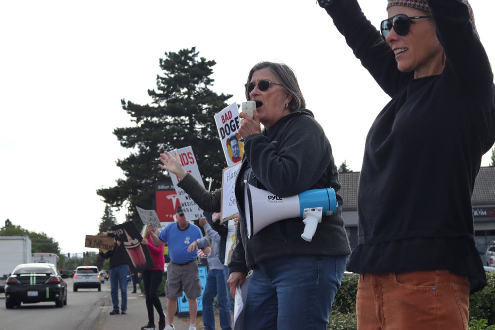 A group of protestors hold picket signs and wave along the sidewalk next to the road. One visible picket sign reads, “BAD DOGE”, and depicts an image of Elon Musk. The sign for the Tesla dealership, which depicts a white Tesla logo on a red background, can be seen behind the protestors.

At 5:00pm on May 14th, 2025, protestors gathered outside the Vancouver, WA Tesla dealership to protest the fascist regime of US President Donald Trump and unelected billionaire oligarch Elon Musk. 

