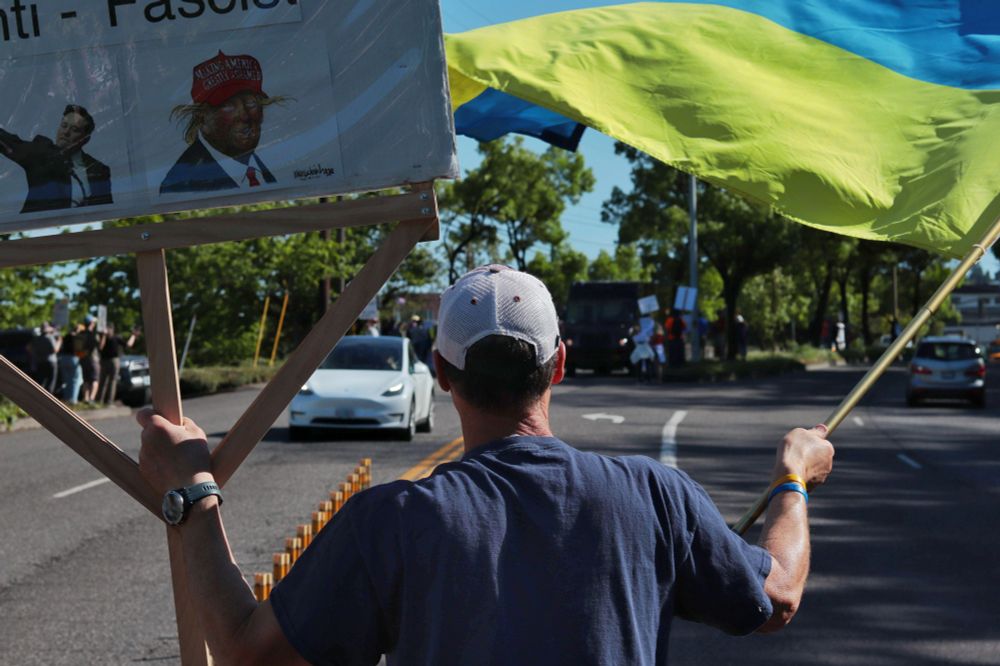 A close-up image of the back of a protestor who stands on a median in the middle of the road. The protestor holds a sign while waving a full-size Ukrainian flag. The sign depicts an image of President Donald Trump underneath the word, “FASCIST”. Past the protestor, a white Tesla drives on the road toward the viewer. 

At 3:45pm on May 5th, 2025, protestors gathered outside the Portland Tesla dealership on Macadam Ave to protest the fascist regime of Donald Trump and Elon Musk.
