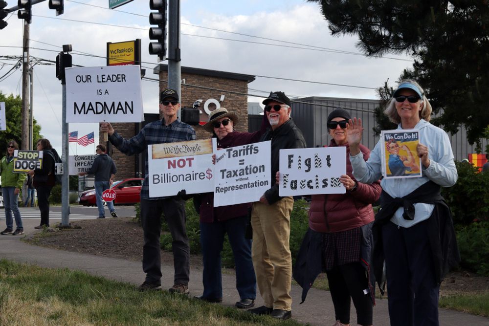 Five protestors on the sidewalk hold picket signs while looking at the viewer. Some of the protestors are smiling and waving. From left to right, the signs read: “OUR LEADER IS A MADMAN”, “WE THE PEOPLE | NOT | Billionaire$”, “(Trump’s) Tariffs = Taxation without Representation”, “Fight Fascism”, and “We Can Do It!”. The sign furthest to the right depicts an image of Rosie the Riveter. 

At 5:00pm on May 14th, 2025, protestors gathered outside the Vancouver, WA Tesla dealership to protest the fascist regime of US President Donald Trump and unelected billionaire oligarch Elon Musk. 
