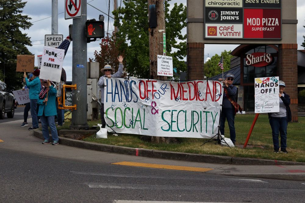 Demonstrators hold up a banner at one corner of the intersection. The banner reads "HANDS OFF OUR MEDICAID & SOCIAL SECURITY." Around the banner, several other demonstrators carry picket signs.

At 4pm on May 19th, 2025, demonstrators in Vancouver, WA gathered at the intersection of Mill Plain and Chaklov to protest the destruction of essential social services by US President Donald Trump's fascist regime.