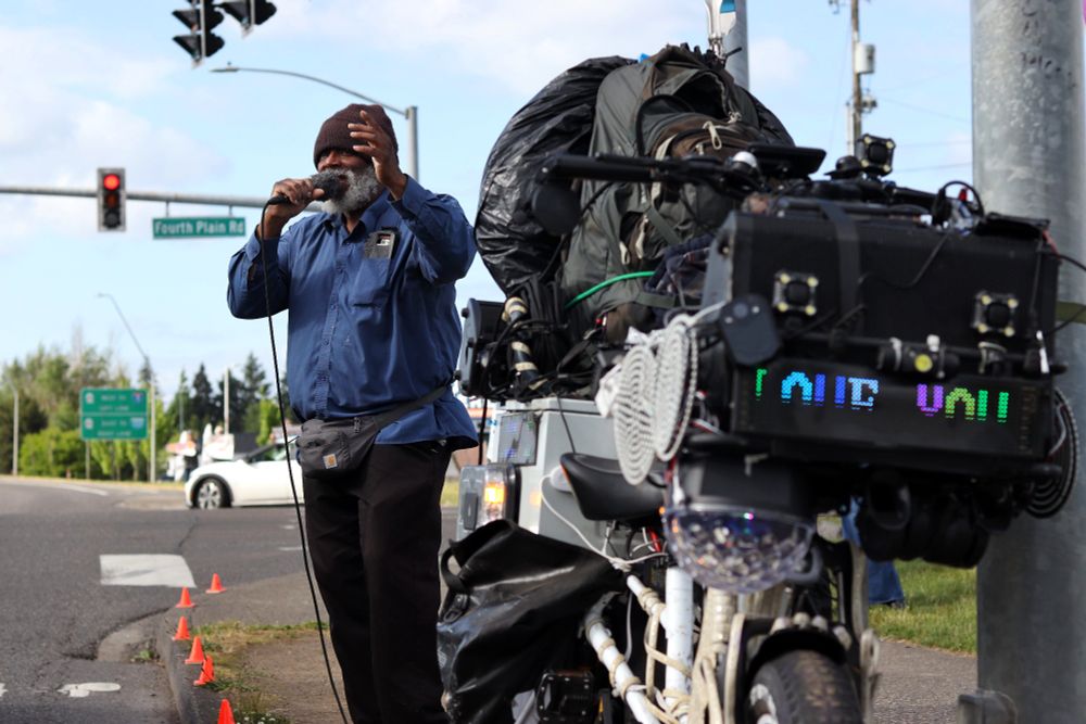 A community member on the sidewalk holds a microphone singing. To their right, the community member’s bicycle is propped up. Multiple bags and belongings are strapped to the bicycle. An LED sign on the front of the bike displays the words, “LOVE YOU”. 

At 5:00pm on May 14th, 2025, protestors gathered outside the Vancouver, WA Tesla dealership to protest the fascist regime of US President Donald Trump and unelected billionaire oligarch Elon Musk. 
