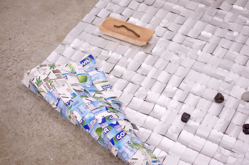 A sheet made from small rectangular fragments of recycled, metallic, tetrapak packaging material held together by tiny stainless steel jump rings rests on a concrete floor. Various sculptural objects rest on its surface: a hand-carved bowl holds water and a floating piece of driftwood, chunks of petrified wood and a single smooth, oblong stone are cast like dice. 