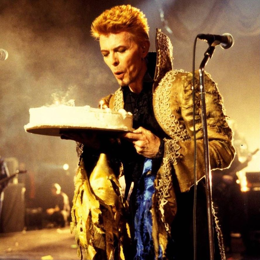 David Bowie resplendent in gold holds a birthday cake and is blowing on the candles.