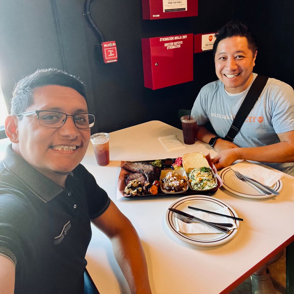 Two people are sitting at a small restaurant table with a platter of assorted barbecue dishes, including smoked meats, sides, and bread, placed between them. Each person has a drink with ice and a plate with utensils. The setting has a casual, modern vibe with dark walls, visible fire alarm boxes, and natural daylight coming in from the left. One person is wearing a black collared shirt, and the other is wearing a light gray T-shirt with a black shoulder strap.
