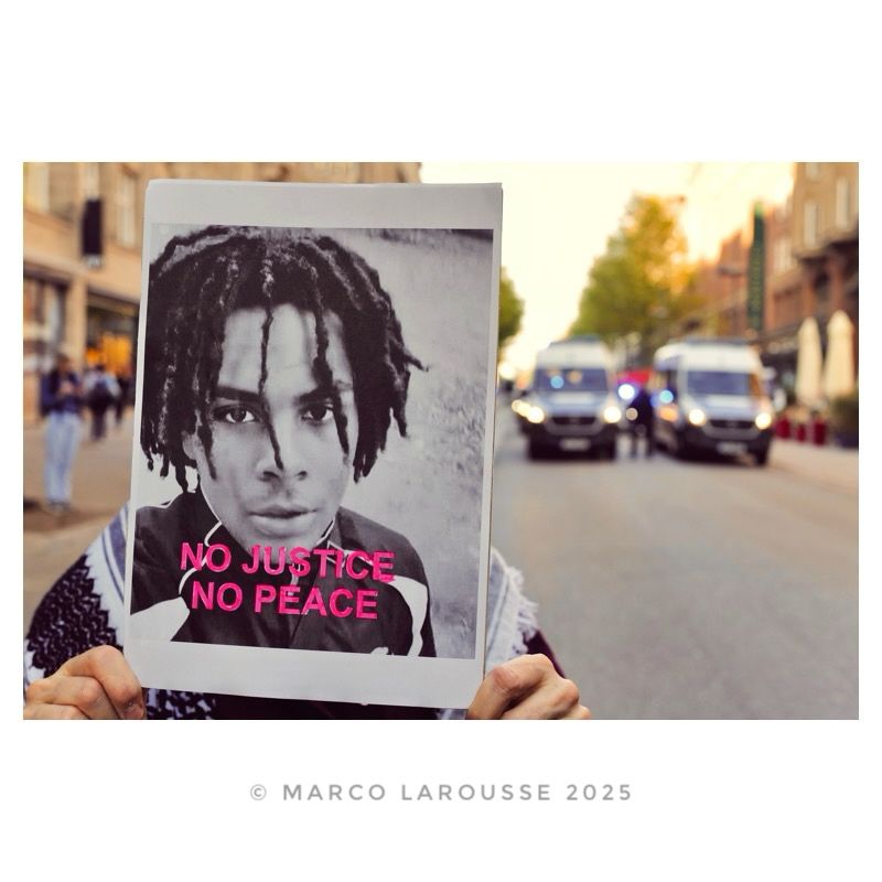 Am hinteren Ende des Demozugs hält eine Frau ein Schild mit dem Foto von Lorenz auf dem steht: „No Justice - No Peace“. Dahinter fahren ca. ein Dutzend Polizeifahrzeuge.