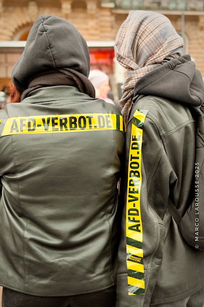 Zwei Demonstranten haben gelb-schwarze Warnstreifen auf ihren Jacken befestigt, auf denen steht: „AFD-VERBOT.DE“