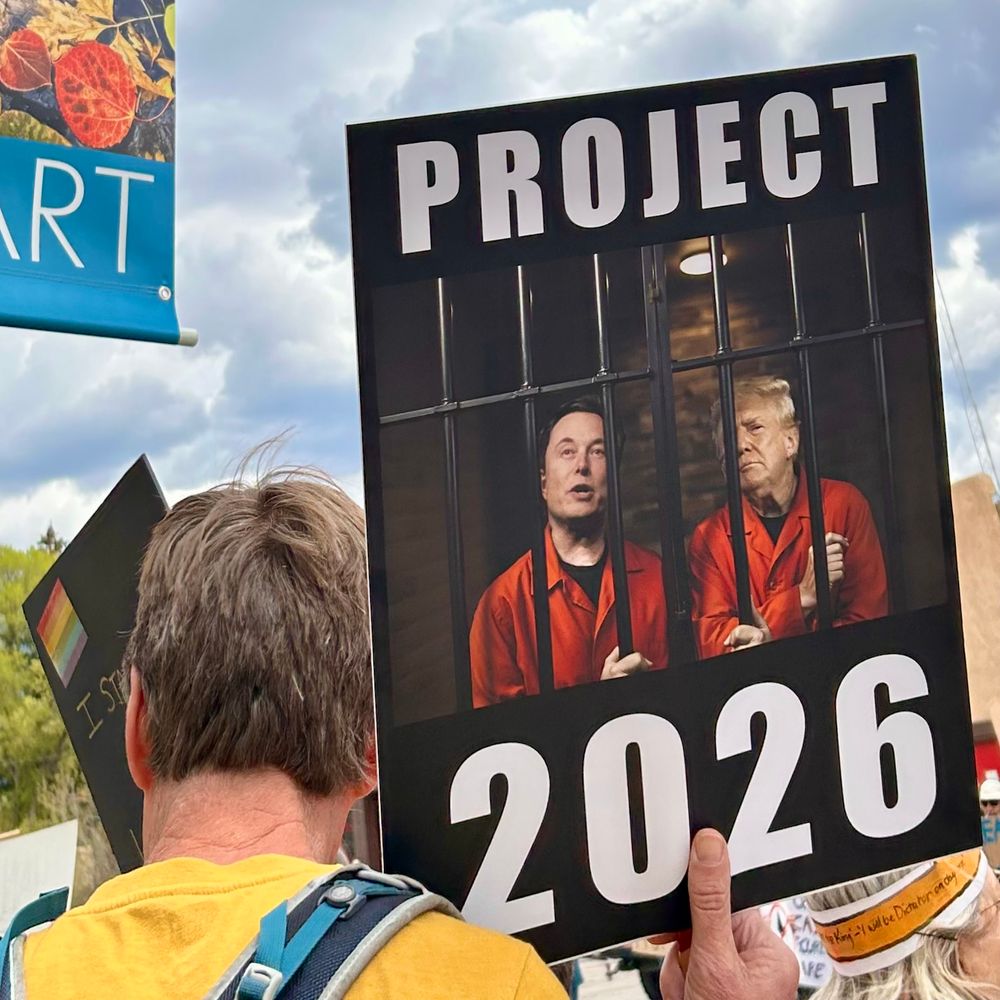 Protestor holding sign, Elon Musk and Donald Trump in orange jumpsuits behind prison bars with dark background, words “Project 2026”