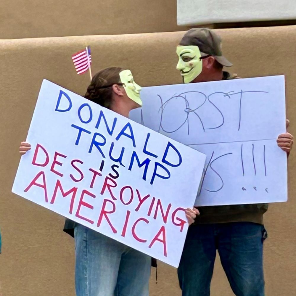 50501 Hands Off protest today in Taos, New Mexico, two protestors wearing Guy Fawkes masks, holding sign “Donald Trump is Destroying America”