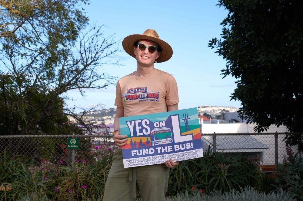 Aidan in a Fund the Bus t-shirt, sunglasses and sun hat holding a "Yes on L, Fund the Bus" window sign in a garden with a San Francisco landscape in the background