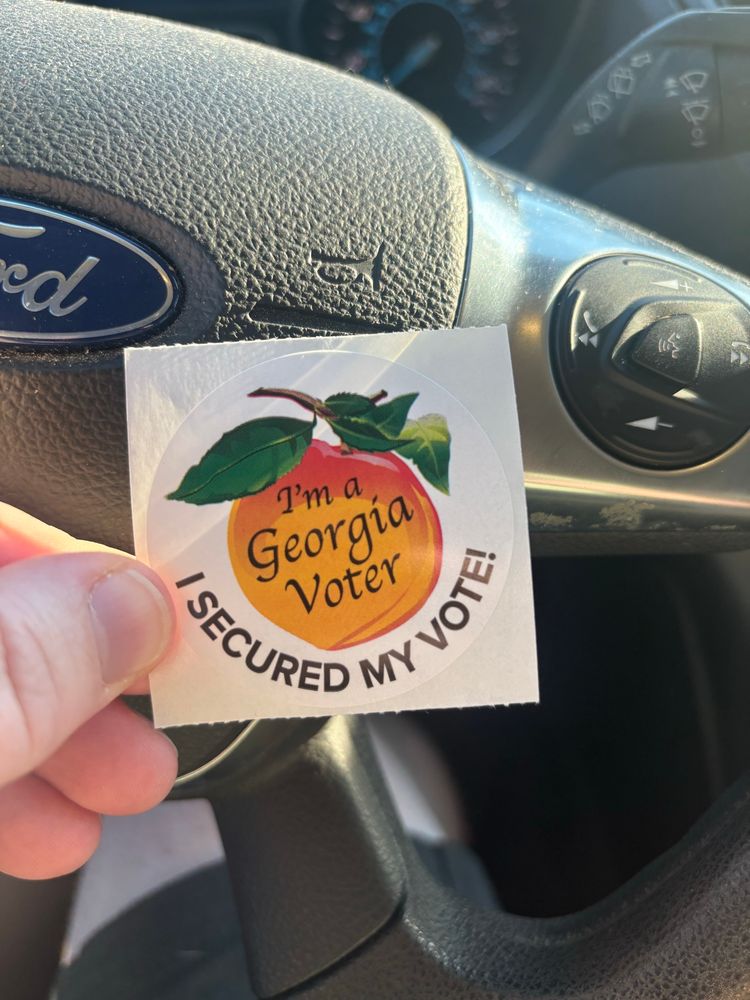 A sticker from the Georgia polls depicting an orange and red peach with a stem and green leaves on a white background. On top of the peach black letters read "I'm a Georgia voter" and underneath the peach it says "I secured my vote!"