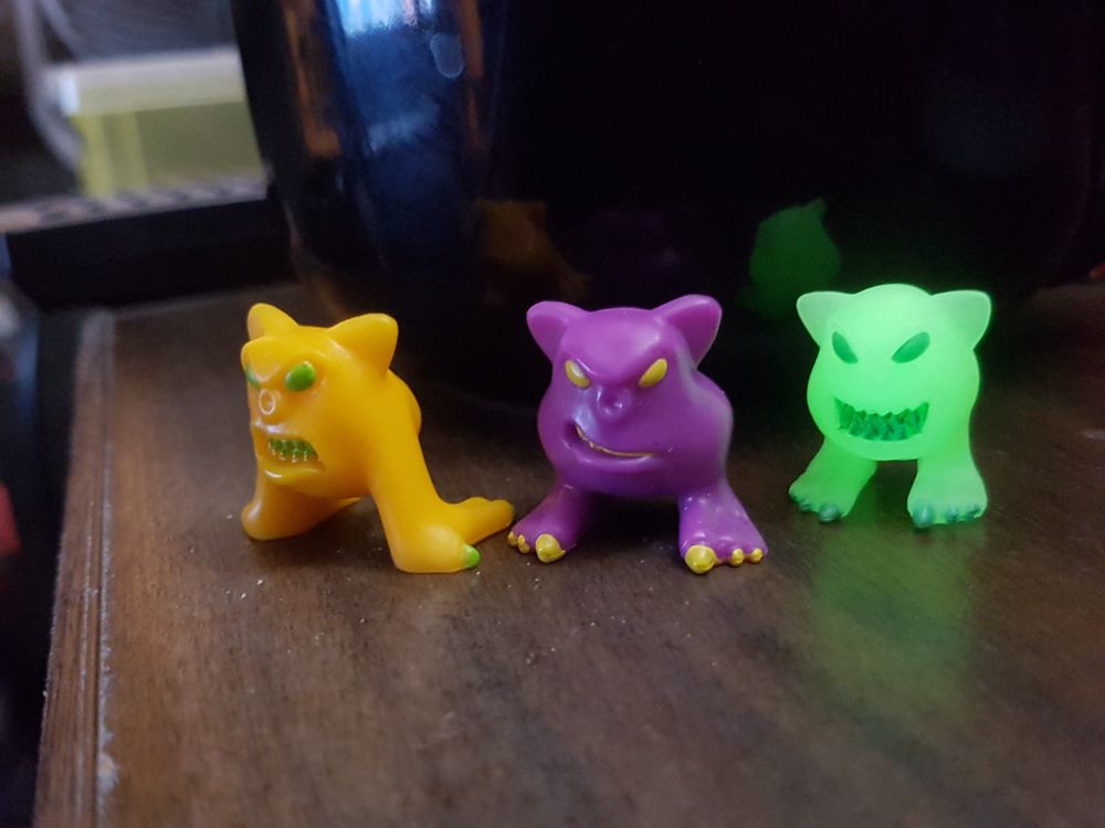 Three Mordles, a yellow one, a purple one, and a glowing one, stood on a wooden coffee trable, infront of a coffee mug.