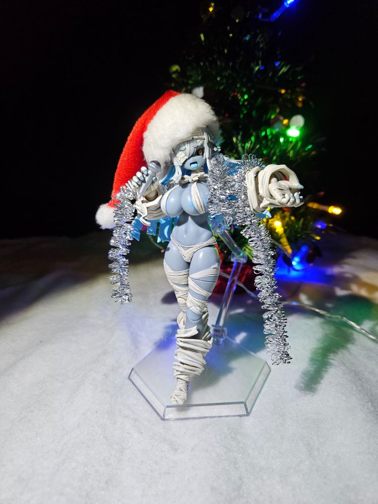 Zombie Grisa wearing garland and a Santa hat in front of a lit up Christmas tree while singing into a microphone