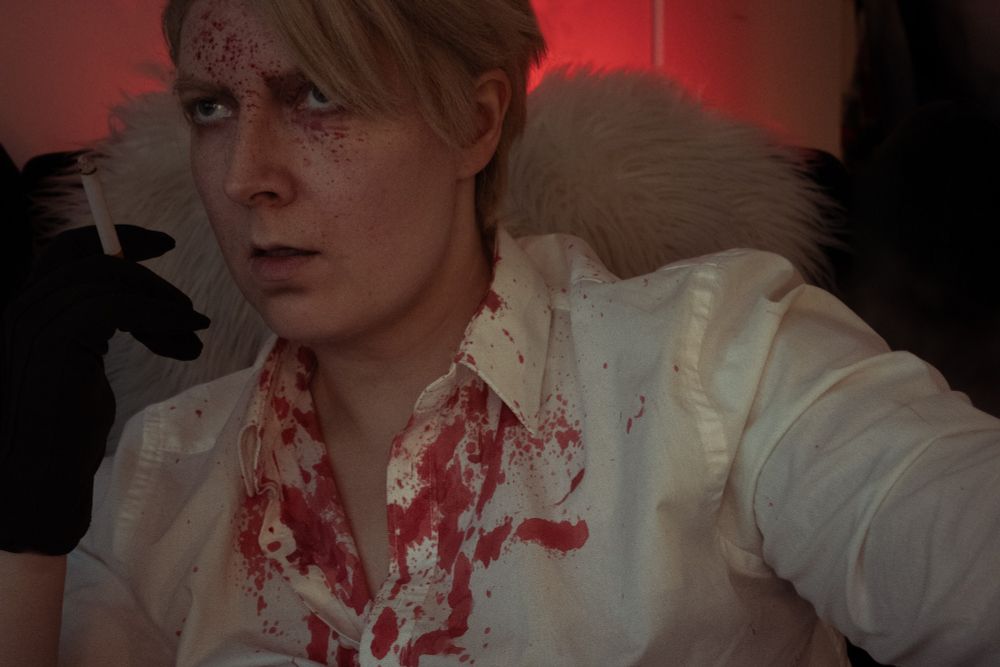 Cosplay of Caesar from Roses and Champagne. He is sitting on a chair in a dark room and wearing a shirt with red stuff. He looks little bit angry or annoyed 