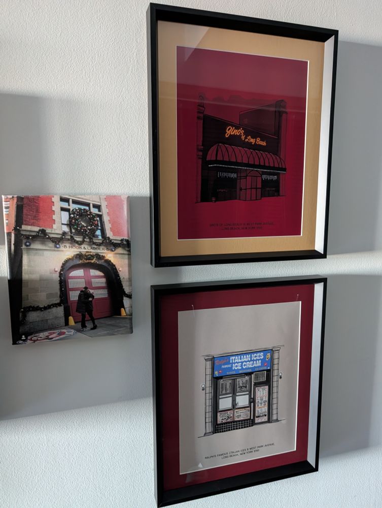
An angled view of three framed art pieces hanging on a white wall. The top frame, with a red background and a light beige border, contains an illustration by artist Henry Kaye of Gino's of Long Beach. Below it is a second frame, with a red mat and a thin white border, holding another Henry Kaye illustration of Ralph's Famous Italian Ices. To the left of the two illustrations, a third, smaller photo print is mounted on the wall. It shows the front of a red brick building with large red doors and holiday wreaths. It is the Ghostbusters Firehouse in New York, New York. Patrick and Kara are embracing in front of the doors.