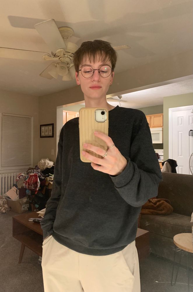 Mirror selfie of ash in a dark grey sweatshirt with light tan sweatpants. They are wearing thin-rimmed black glasses and their hair is cut short with a choppy, textured fringe covering their forehead.