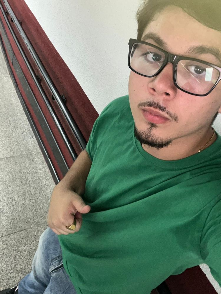 Theo hj, um homem branco, cavanhaque, óculos, camiseta verde e calça jeans fazendo joia com o dedo 