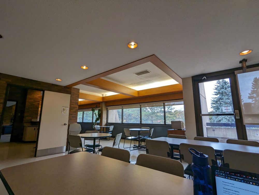 Wide-angle photo of an empty study area/lounge with tables, chairs, a small kitchenette, and large windows on the far wall.