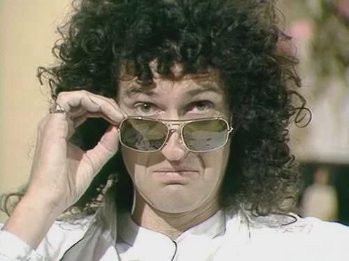 Image of Queen guitarist Brian May making a sour expression while peering over the sunglasses perched atop his nose. 
