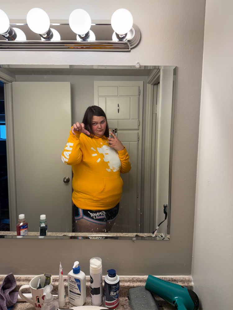 A reflected selfie of Kit in a bathroom mirror, wearing a yellow hoodie with a picture of a hyena laughing. The sleeves say “hahaha”