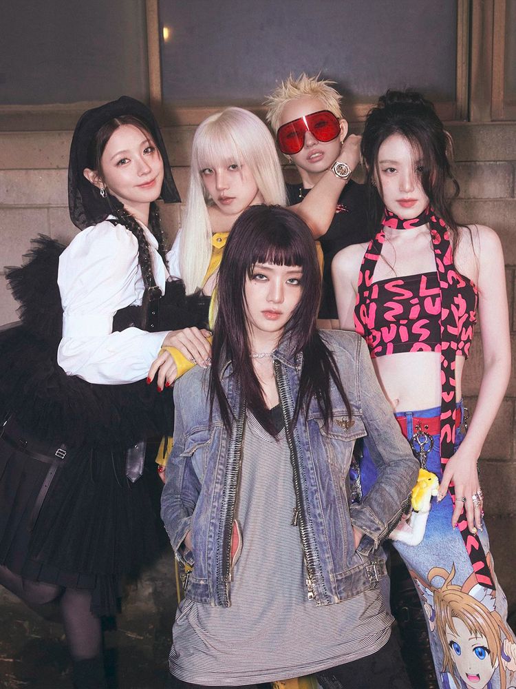 I-DLE members grouped together, looking at the camera, standing in front of a concrete wall at night