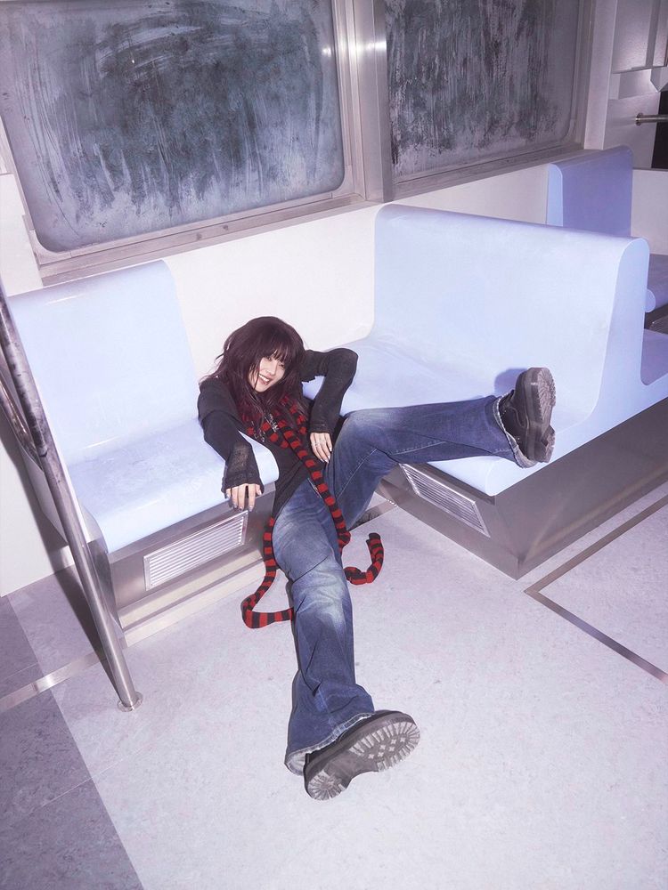 Minnie is playfully sitting on the floor between two benches in a subway car. She is wearing an urban outfit with a red and black striped scarf