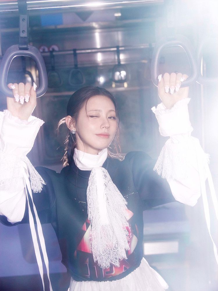 Close-up photo of Miyeon, inside a subway car holding the hand straps. She is wearing a white lace dress with ruffled sleeves and a black jacket