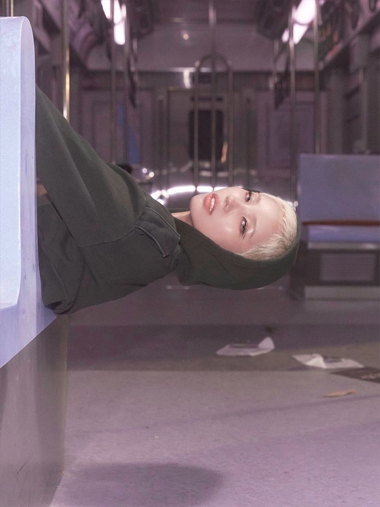 Soyeon, with short blonde hair and wearing a dark green hood, is lying on a bench inside an empty subway car, staring at the camera