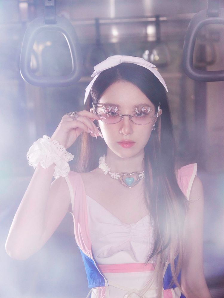 A close-up photo of Shuhua inside the subway car, wearing pink-tinted glasses and a white headband. Her upper Sailor Moon-inspired outfit is visible, along with a heart-shaped choker