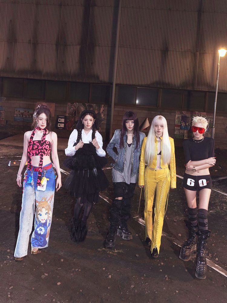 I-DLE members standing in a line, gazing at the camera, they are on an empty parking lot at night, with rocks scattered across the ground