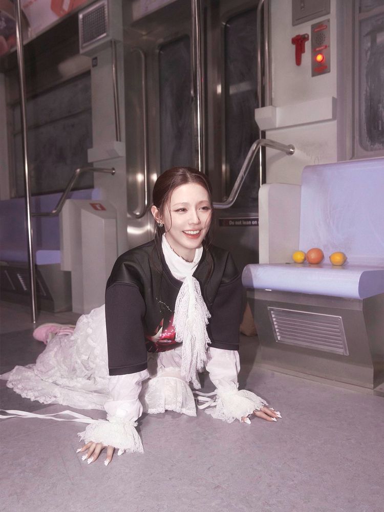 Miyeon is wearing a white lace dress with ruffled sleeves and a black jacket. She kneels, with both hands down, on the subway car floor and smiles