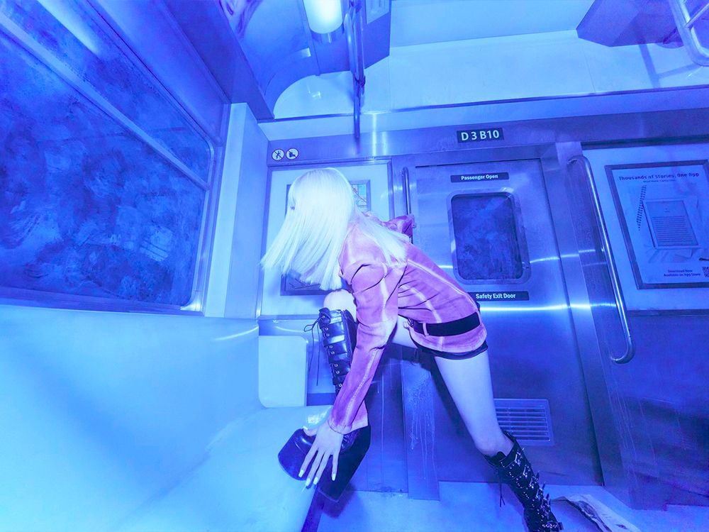Yuqi is inside a blue-lit subway car, with one foot on a bench, striking a pose as if she's about to climb onto it.