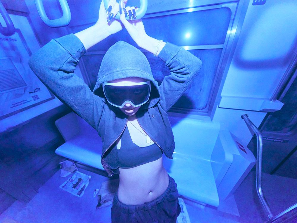 Soyeon inside a blue-lit subway car, she is wearing a dark green hood and oversized black visor sunglasses