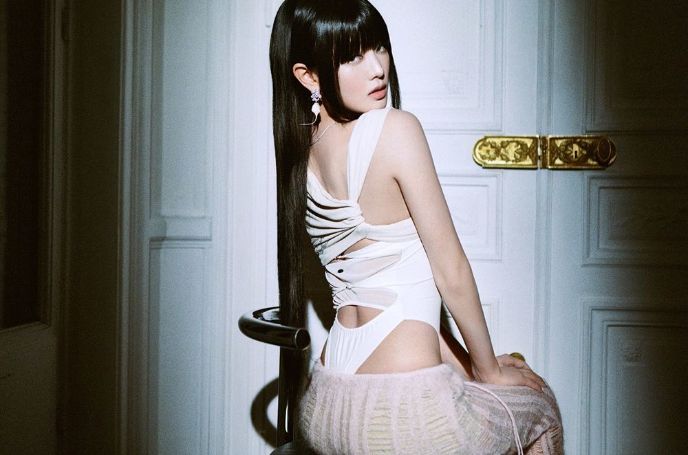 Minnie (i-dle) sitting with her back turned slightly, looking over her shoulder toward the camera
