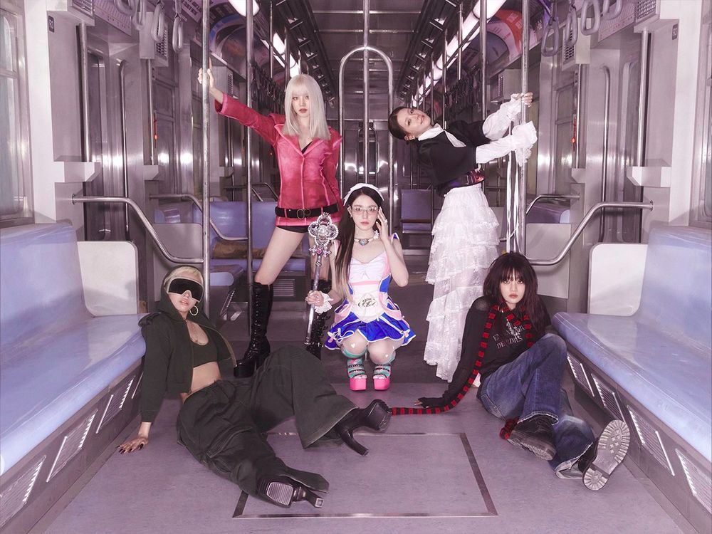 i-dle members pose in an empty subway car. Shuhua is sitting on her heels at the center, flanked by Soyeon and Minnie who are seated on the floor. Yuqi and Miyeon stand behind them, holding onto the metal bars. Shuhua is dressed in a Sailor Moon-inspired outfit and holding a staff, while the other members wear urban street-style clothing