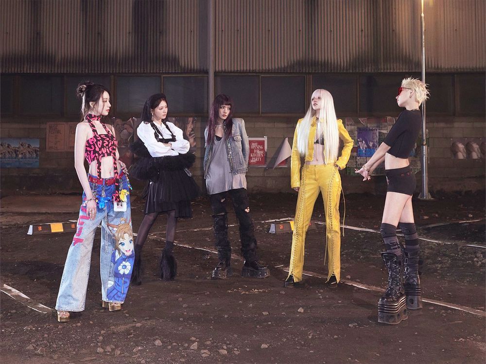 I-DLE members standing in a circle in an empty parking lot at night, with rocks scattered across the ground