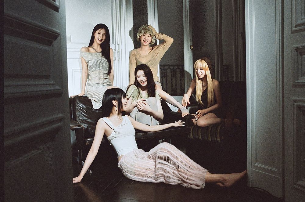 A group photo of i-dle: Minnie is sitting on the floor, Shuhua and Yuqi are sitting on chairs, and Miyeon and Soyeon are standing. They are laughing at each other
