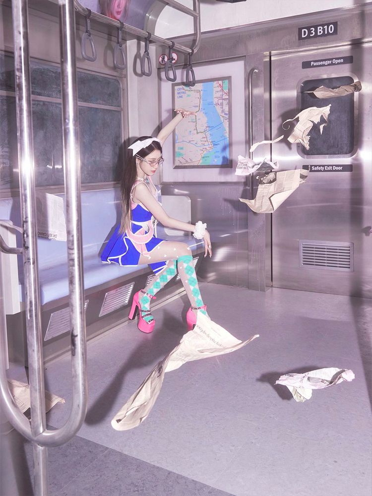 Shuhua sits on a bench inside an empty subway car, dressed in a Sailor Moon-inspired outfit with blue and pink accents. She strikes a magical pose with one hand raised, as loose papers float around her, as if controlled by superpowers