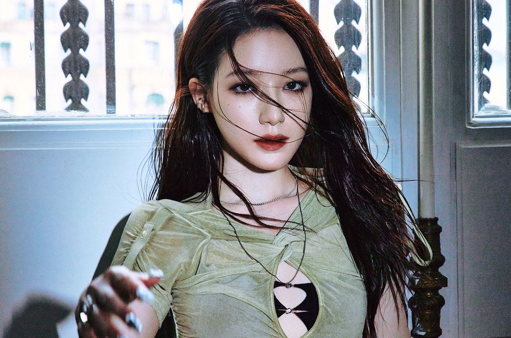 Close-up photo of Shuhua (i-dle) with some hair over her face, one hand slightly moving towards the camera while she gazes into it