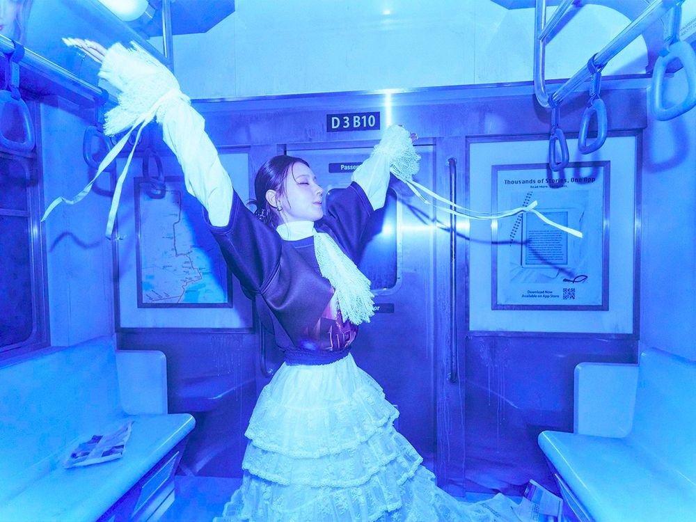 Miyeon, inside a blue-lit subway car, with her eyes shut and hands raised as if she is dancing. She is wearing a white lace dress with ruffled sleeves and a black jacket