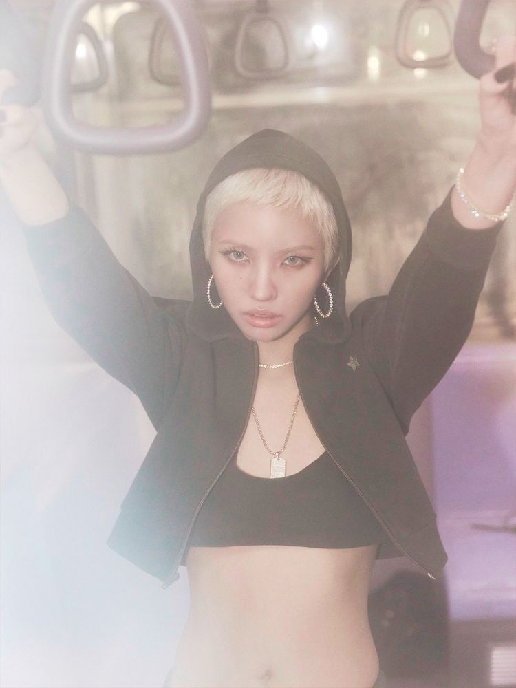 Close-up photo of Soyeon's face and upper body inside a subway car. She has short blonde hair and is wearing a dark green hood