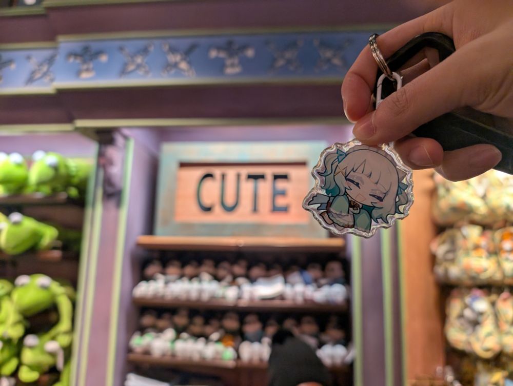 A keychain charm of VTuber Maid Mint Fantome held up against a store shelf with the banner 'CUTE' in the background