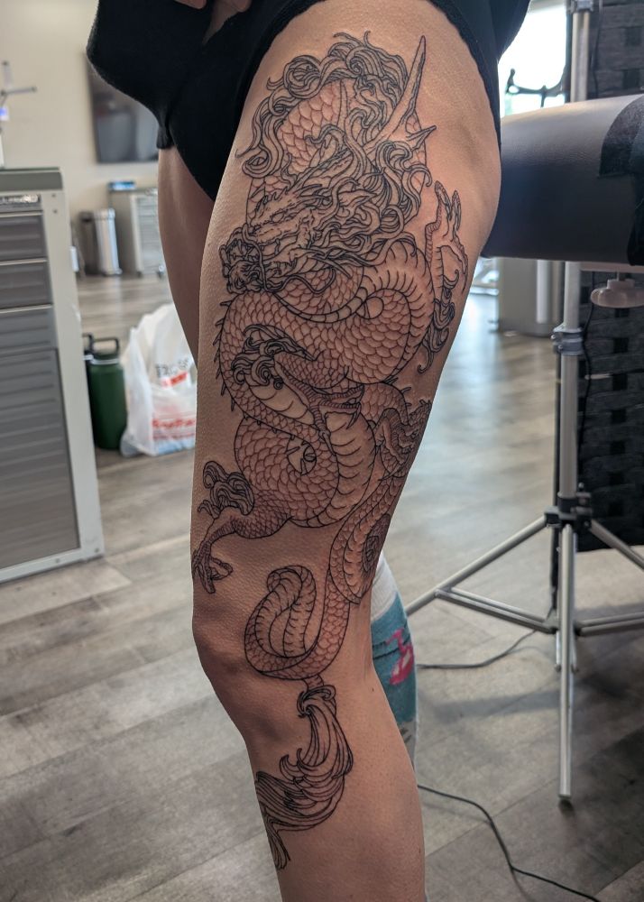 Karin's left thigh. There is a massive black linework Japanese dragon tattooed from just below the 3/4 outside of her hip to midway down the front of her calf, with the tail wrapping around the knee.
