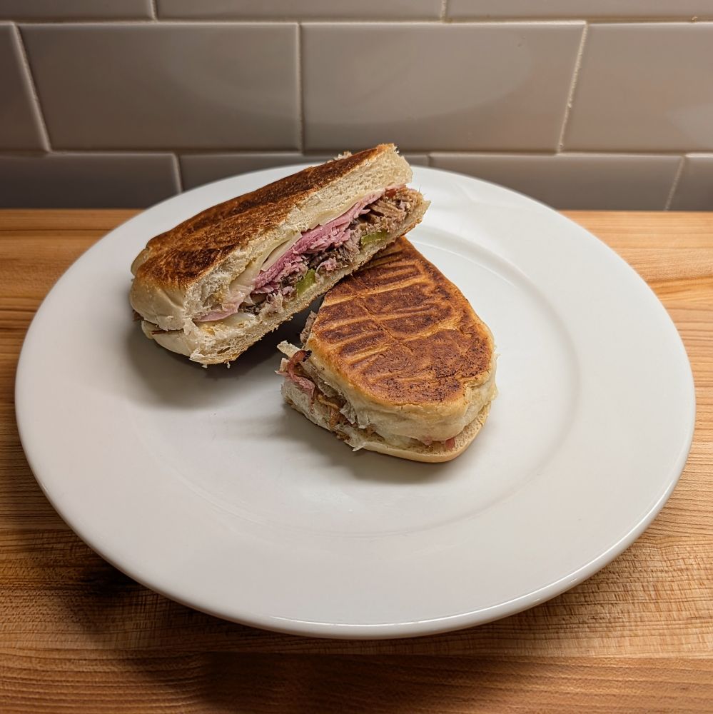 A beautifully pressed and toasted cubano, full of mojo pork, ham, cheese, cornichons, and mustard, sliced in half on a plate
